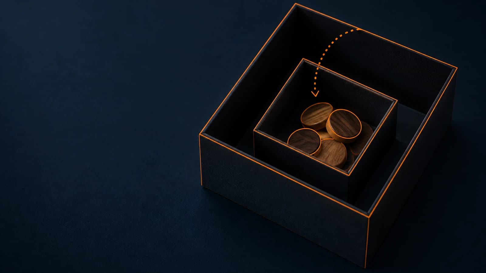 Annotated photograph on a deep navy surface, seen from above at an angle: a small open black box sits inside a larger open black box, holding a handful of round wooden tokens lit by warm light. Thin bright orange lines trace the edges of both boxes, and a dotted orange arrow curves from the larger box down into the smaller one.